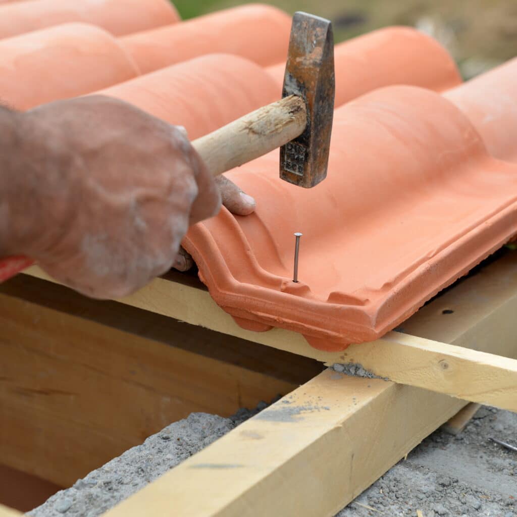 Pose de tuile : un ouvrier fixe une tuile en terre cuite. Ouvrier clouant une tuile en terre cuite avec un marteau sur la charpente en bois. Détail de la pose du toit.